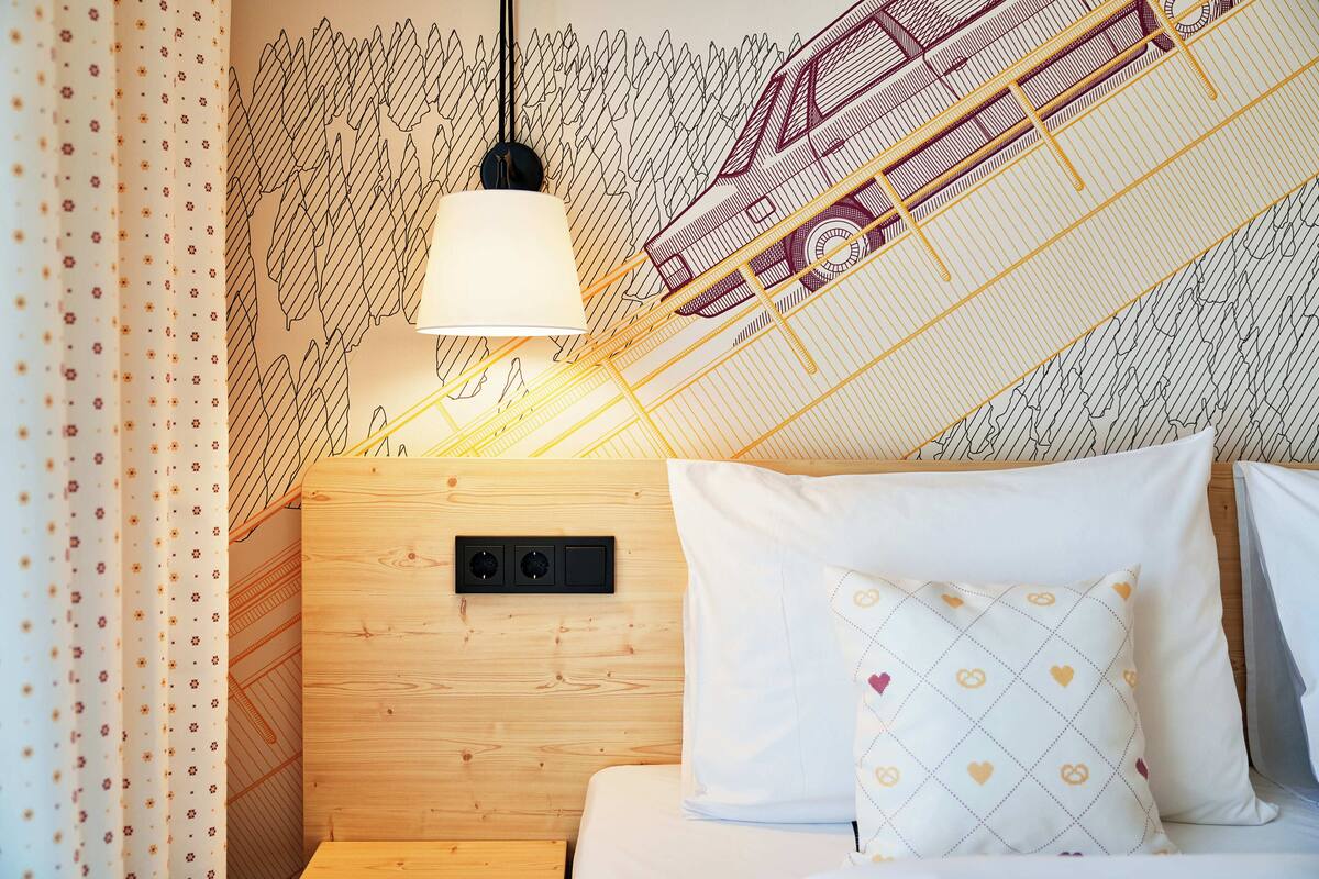 Un dormitorio con cabecera de madera, cama con ropa de cama blanca y almohadas estampadas, lámpara colgante y pared con diseño geométrico.
