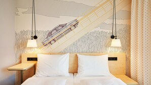 Una cama doble con sábanas blancas, dos mesitas de noche con lámparas y un mural en la pared con un tren.