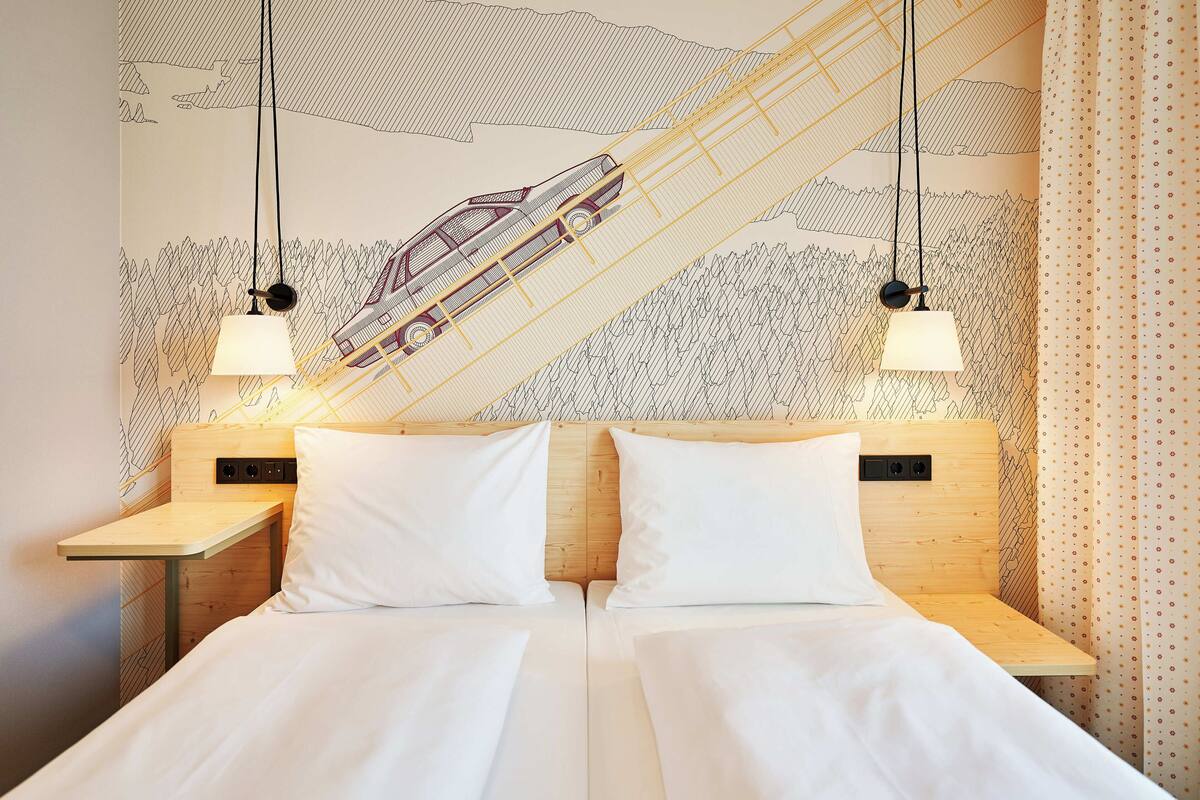Una cama doble con sábanas blancas, dos mesitas de noche con lámparas y un mural en la pared con un tren.