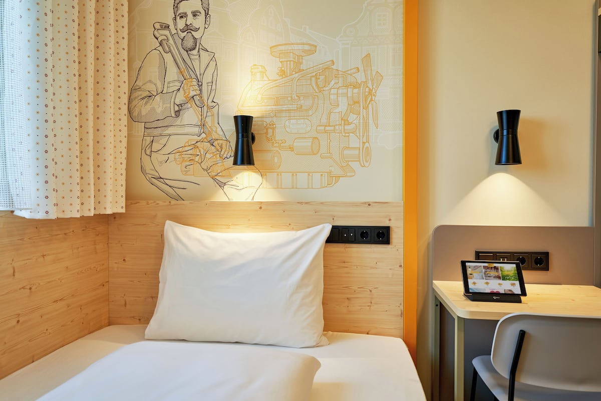 Habitación de hotel con cabecera de madera, cama con ropa blanca, lámpara de pared, pequeña mesa con una tablet y cortina con estampado de lunares.