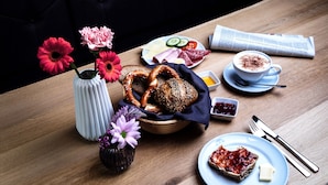 Una mesa de madera con un plato de pan, un bol de pretzels, una taza de café, un periódico y un florero con flores.