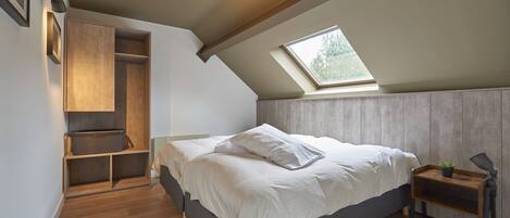 Quarto com teto inclinado, claraboia, cama com roupa de cama branca, guarda-roupa de madeira e mesa de cabeceira com abajur.
