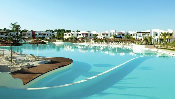 Ein Hotelpool mit Liegestühlen und Sonnenschirmen, umgeben von weißen Gebäuden.