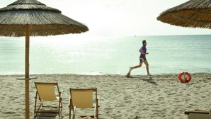 Ein Strand mit zwei Holzstühlen und reetgedeckten Sonnenschirmen, einem Rettungsring und einer Person, die am Ufer entlangläuft.