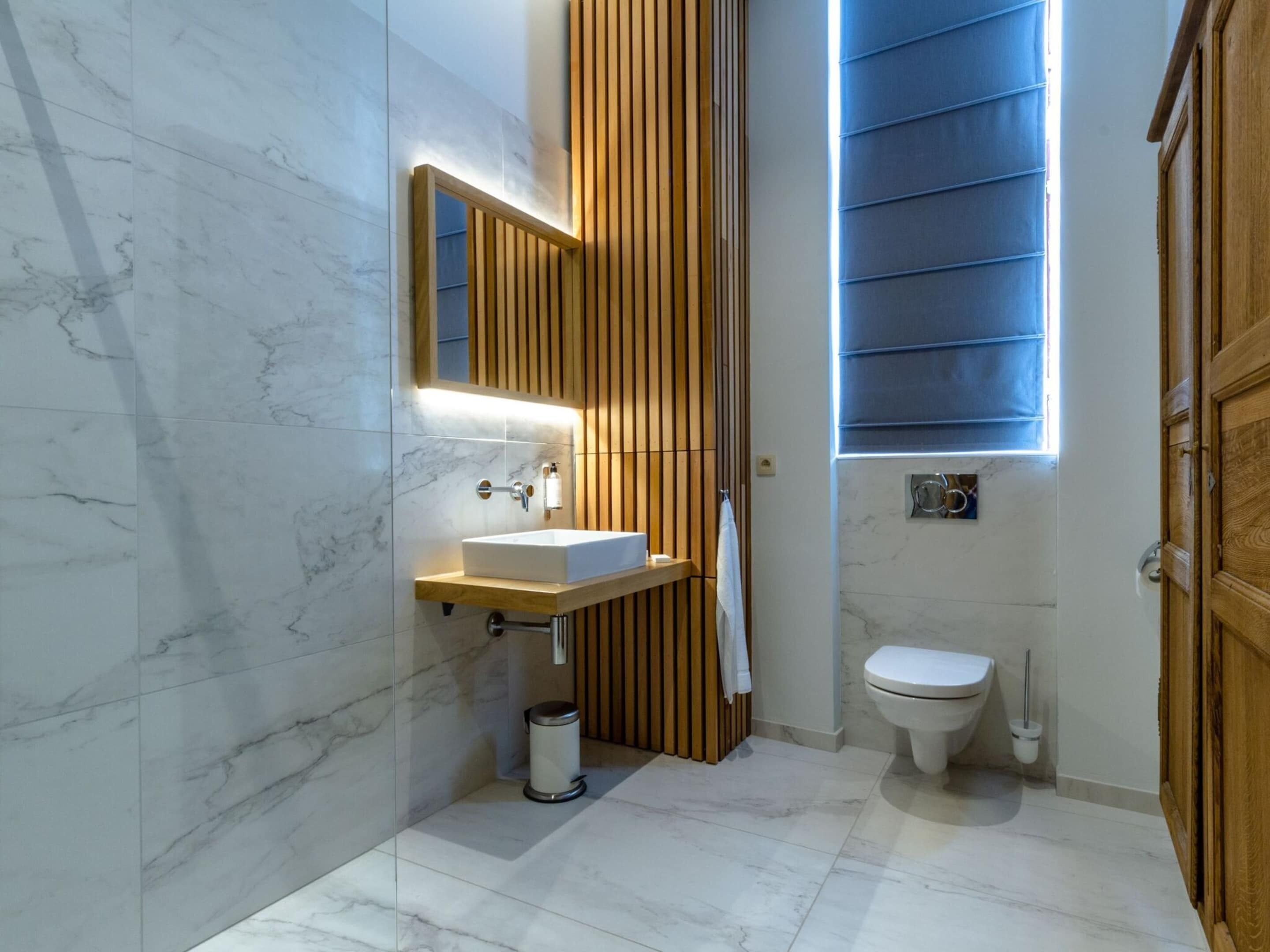 A modern bathroom with a marble floor, a wooden vanity, a white sink, a mirror, a toilet, and a window with blinds.
