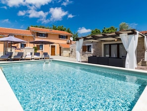 A swimming pool with lounge chairs and umbrellas, a stone building, and a covered seating area.