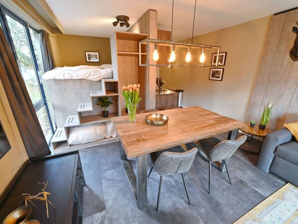 A compact, modern room with a wooden dining table, grey upholstered chairs, a grey sofa, a small kitchen area, and a bed with a view of the outdoors. - Modern Apartment With Terrace (Durbuy)