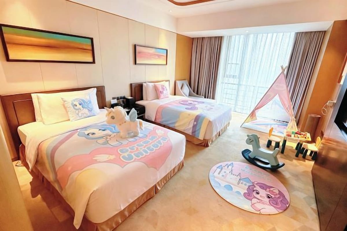 A hotel room with two beds, a TV, a desk, a window with curtains, and a children's play area.