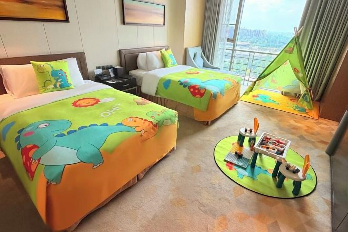 Una camera d'albergo con due letti, ognuno con una coperta a tema dinosauri, un'area giochi con giocattoli e vista sulla città attraverso ampie finestre.