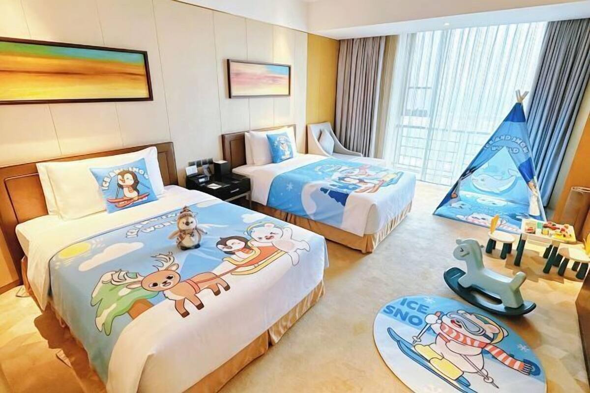 Ein Hotelzimmer mit zwei Betten, einem Schreibtisch, einem Stuhl, einem Fenster mit Vorhängen und einem Spielbereich für Kinder mit Spielzeug.