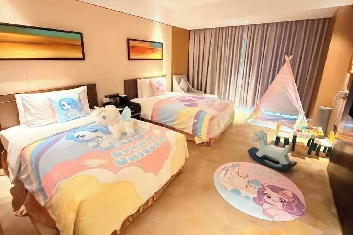 Ein Hotelzimmer mit zwei Betten, jedes mit einer farbenfrohen Bettdecke mit Zeichentrickfiguren. Auf dem Boden steht ein kleines Spielzelt und ein Spielpferd.