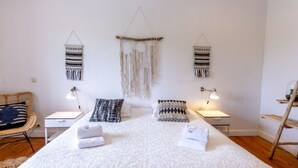 A bedroom with a large bed, two bedside tables with lamps, a macrame wall hanging, and a woven chair.