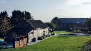 A rustic wooden building with a small porch, a grassy lawn, and a large brick building in the background.