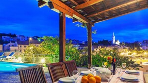An outdoor dining area with a table set for four, overlooking a pool and a scenic view of a town with a church tower. - Villa in Sumartin Near Beach & Private Pool (Selca)