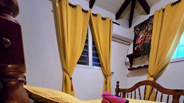 A bedroom with a bed, yellow curtains, a wooden headboard, a bedside lamp, and a wall-mounted shelf with a plant. - Bungalow 2 bdrms in quiet flowered park 3 min from beach and 5 min from town (Deshaies)
