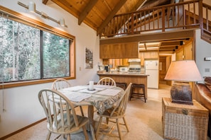 A dining area with a wooden table and chairs, a kitchen in the background, and a staircase leading to an upper level.