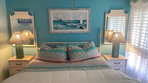 A bedroom with a bed, two bedside tables, a nightstand, a wall-mounted painting, and a window with curtains. - Escape to Classic Anna Maria Island Charm (Bradenton Beach)