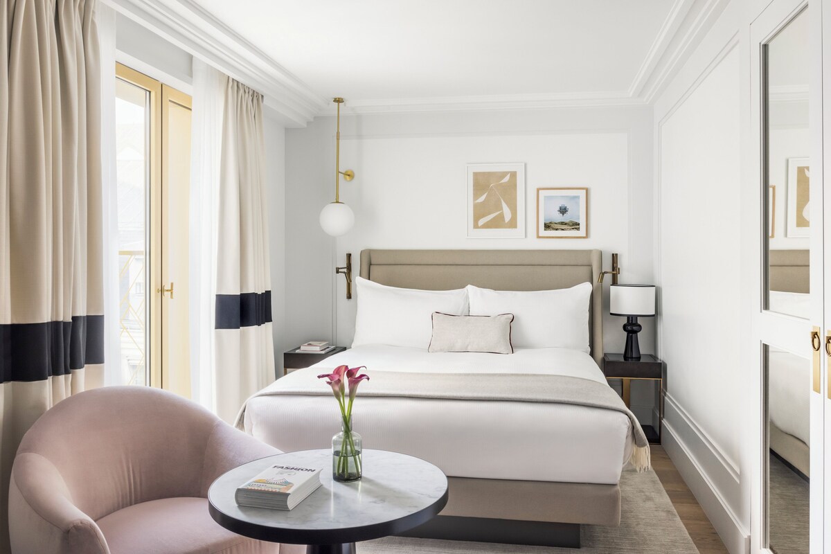 A hotel room with a large bed, a round table with a book and vase, a chair, and a nightstand with a lamp.