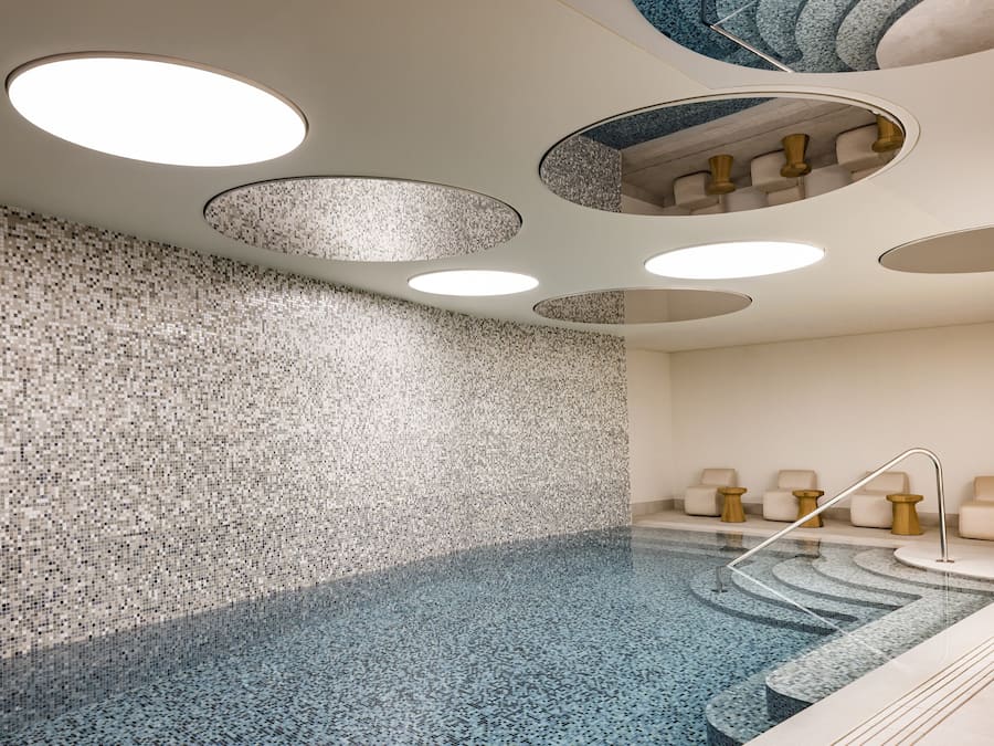 Ein moderner Poolbereich im Innenbereich mit einer Mosaikwand, mehreren runden Deckenleuchten und einer Sitzecke mit Stühlen und kleinen Tischen.