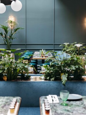 A restaurant with a modern interior, featuring a long dining table with chairs, potted plants, and a bar area.