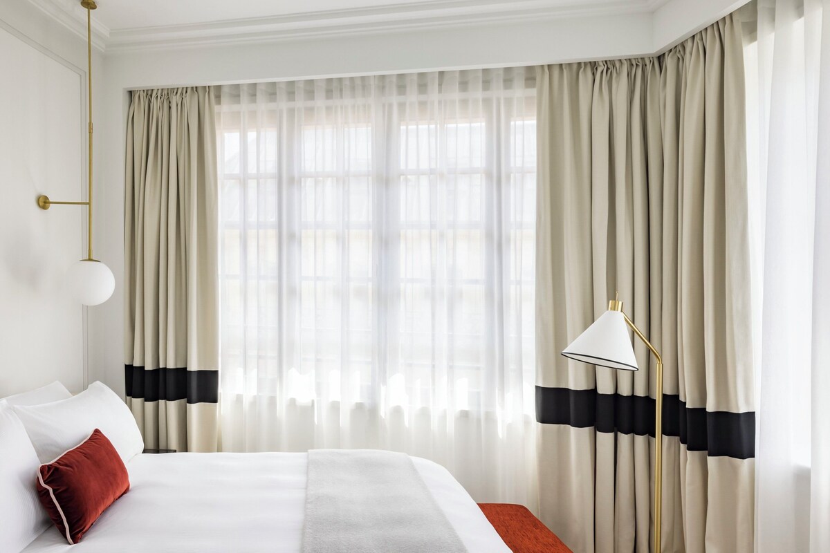A hotel room with a large bed, a bedside lamp, and sheer curtains with black horizontal stripes.