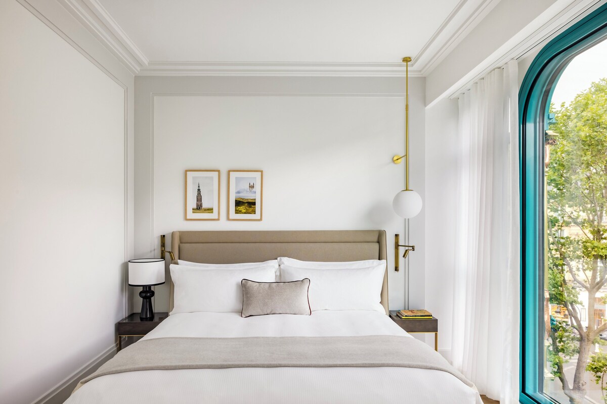 A neatly arranged hotel room with a large bed, a bedside table with a lamp, framed pictures on the wall, and a view through a window.