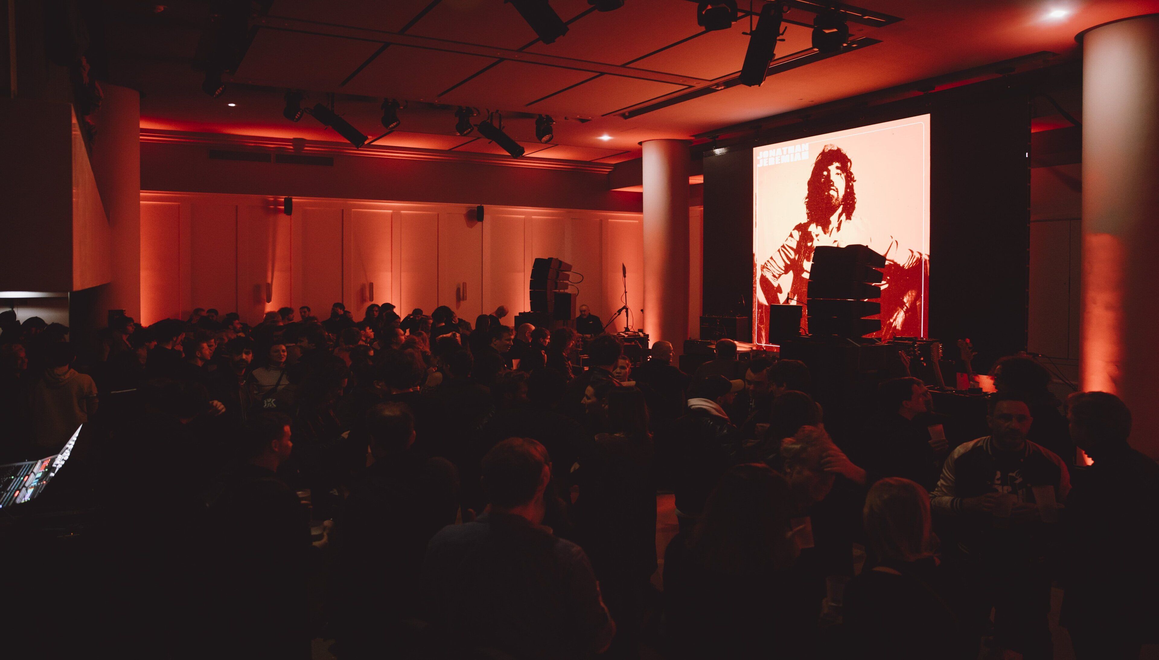 A crowded indoor event with a stage and a large screen displaying a performer.