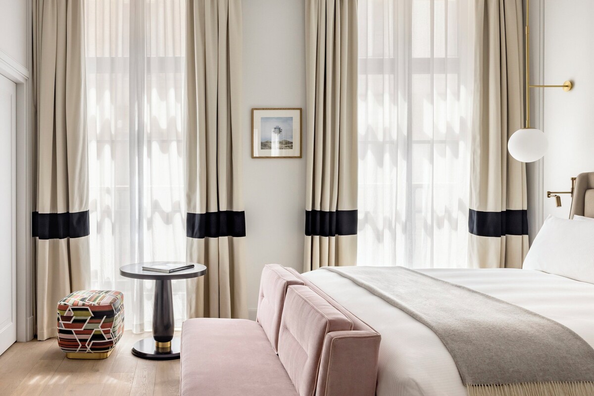 A hotel room with a bed, a small round table, a bench, and curtains.