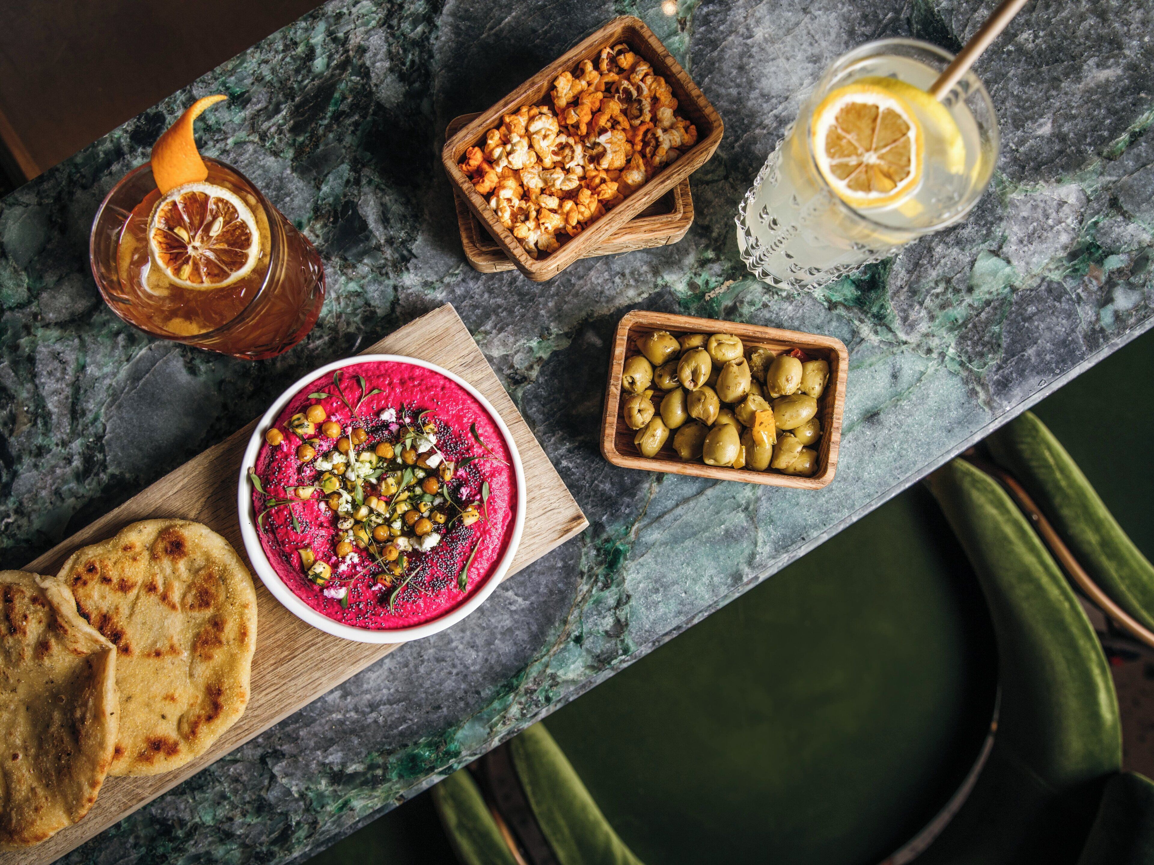 Une table est dressée avec divers aliments et boissons, notamment un saladier de salade de betteraves, un bol d’olives, un bol de pop-corn et deux verres d’une boisson au citron.