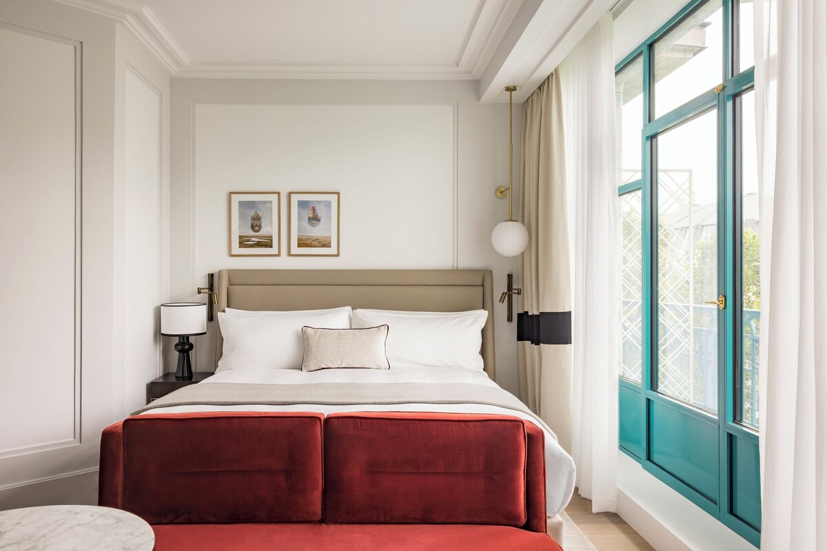 A hotel room with a large bed, a nightstand with a lamp, two framed pictures on the wall, and a window with curtains.