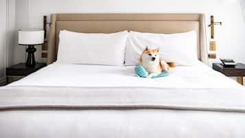 Una cama bien hecha con sábanas blancas, un perro acostado en ella, y una mesita de noche con una lámpara y un teléfono.