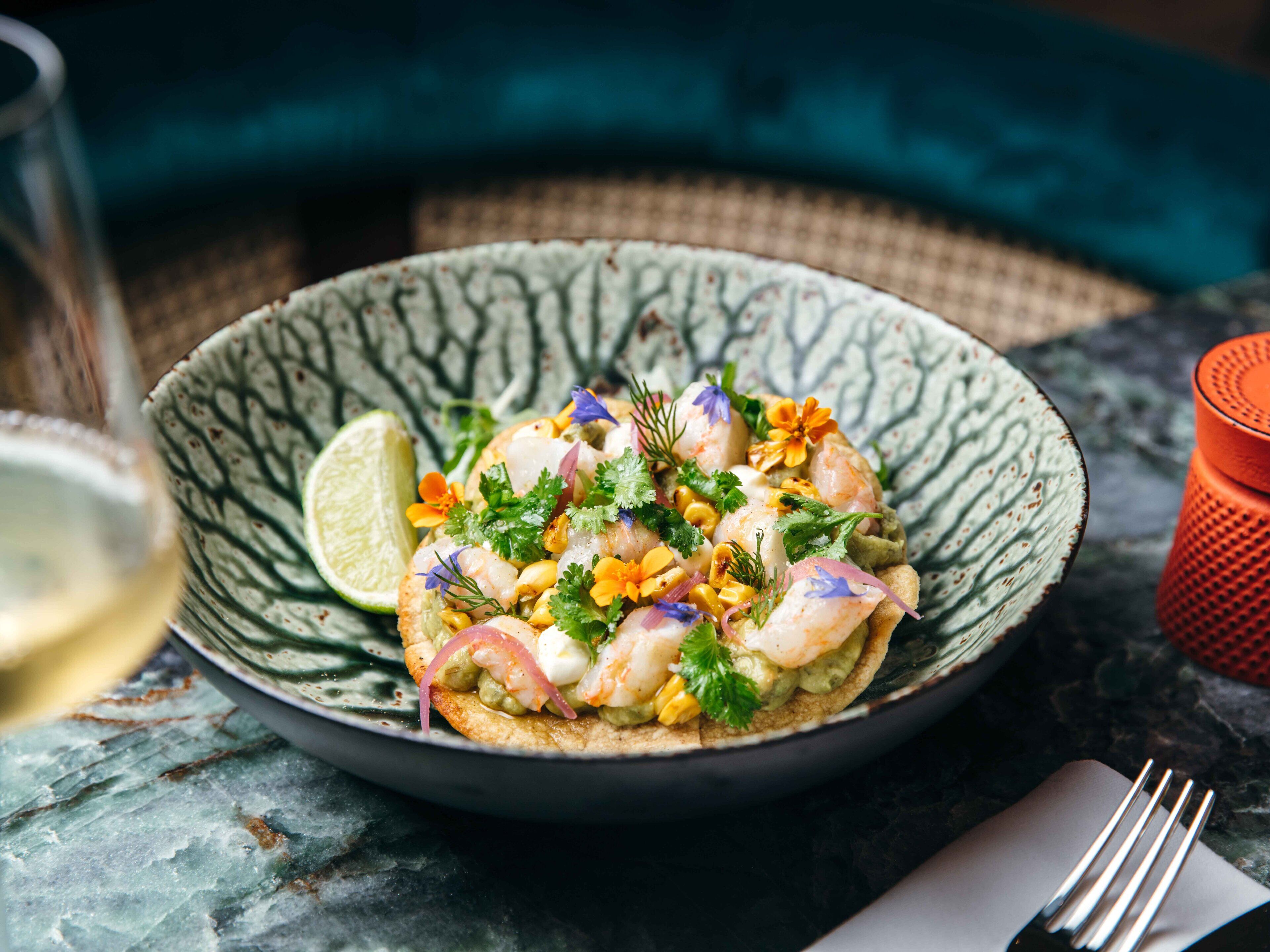 A bowl of seafood salad with a lime wedge, garnished with herbs and flowers, on a textured plate.