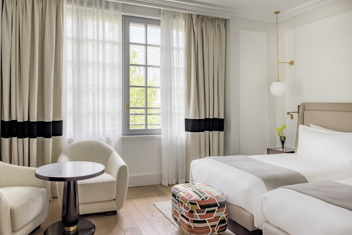 A hotel room with a bed, a chair, a small table, and a window with curtains.