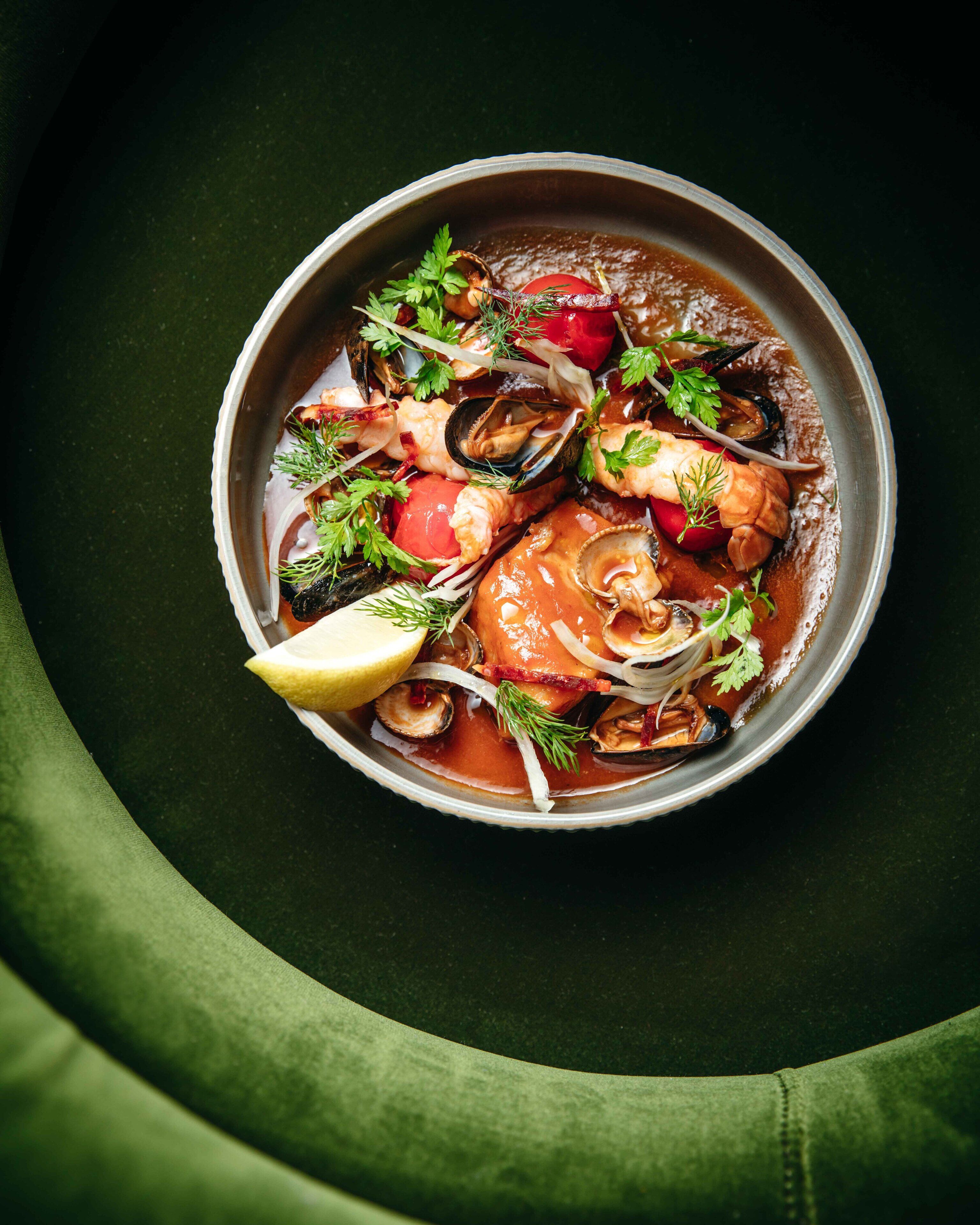 Un tazón de sopa de mariscos con diversos ingredientes como mejillones, tomates y hierbas, servida sobre un plato oscuro con un paño verde debajo.
