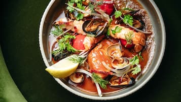 Un tazón de sopa de mariscos con diversos ingredientes como mejillones, tomates y hierbas, servida sobre un plato oscuro con un paño verde debajo.