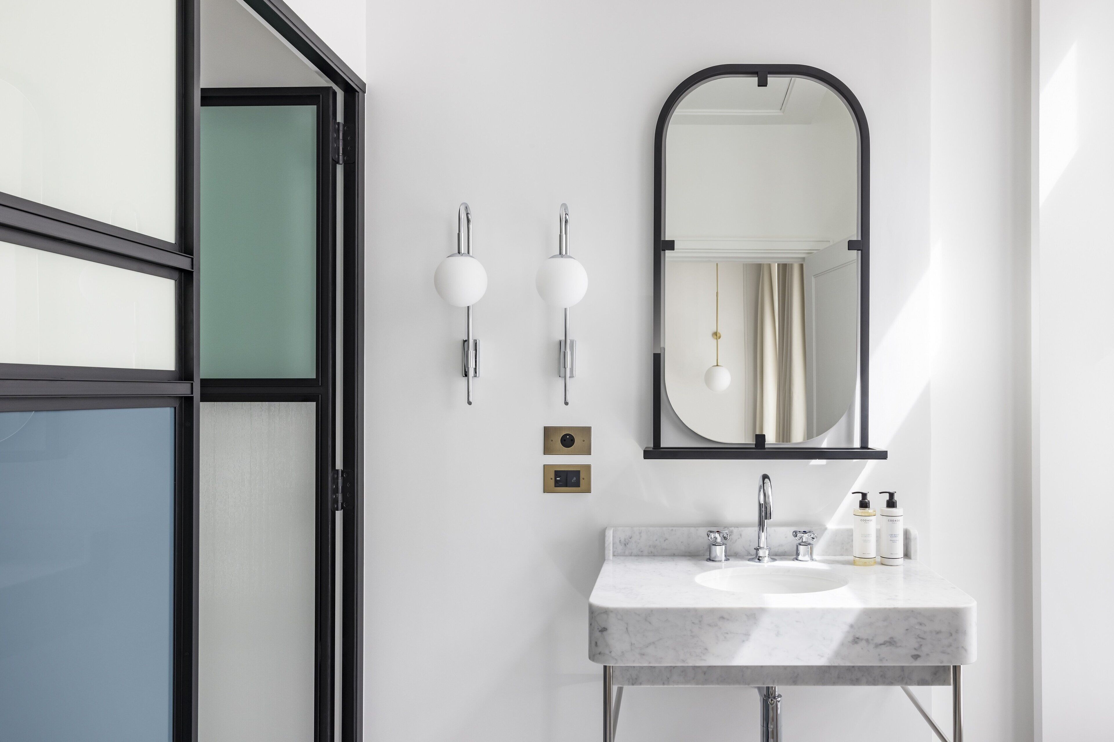 A modern bathroom with a marble sink, two wall-mounted lights, a mirror, and a door with frosted glass panels.
