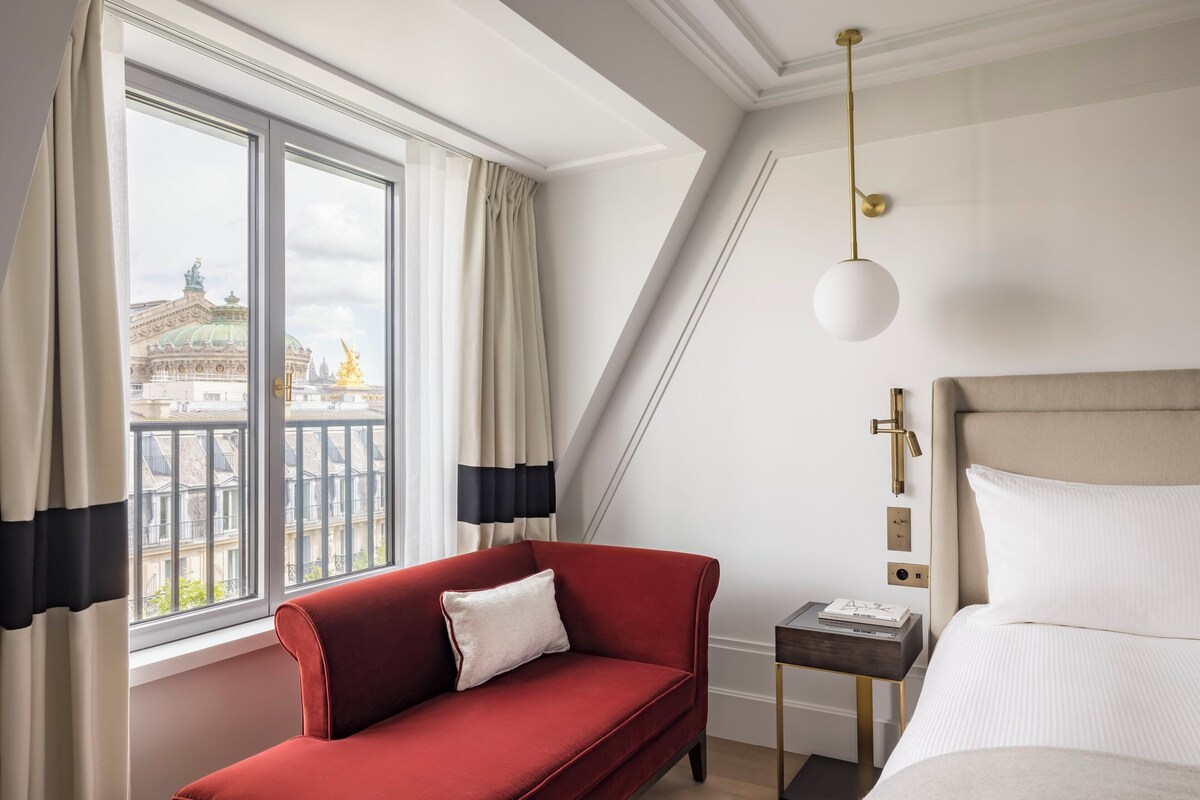 A hotel room with a red sofa, a bed with white bedding, a nightstand, a lamp, and a view of a building through the window.