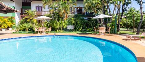 A swimming pool with lounge chairs and umbrellas, surrounded by palm trees and a building with a tiled roof.