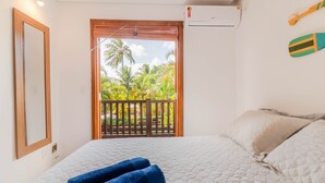 A bedroom with a bed, a window with a view of palm trees, a wooden balcony, and an air conditioning unit. - PF08 Ap Duplex 2 Suites - Praia do Forte (Mata de São João)