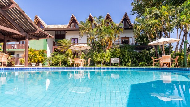 A swimming pool with tiled edges, surrounded by lounge chairs and umbrellas, with a building featuring a tiled roof and palm trees in the background. - PF08 Ap Duplex 2 Suites - Praia do Forte (Mata de São João)