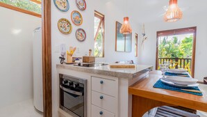 A kitchen with a built-in oven, a sink, and a dining area with a wooden table and chairs. There are decorative plates on the wall and a window with a view of greenery. - PF08 Ap Duplex 2 Suites - Praia do Forte (Mata de São João)