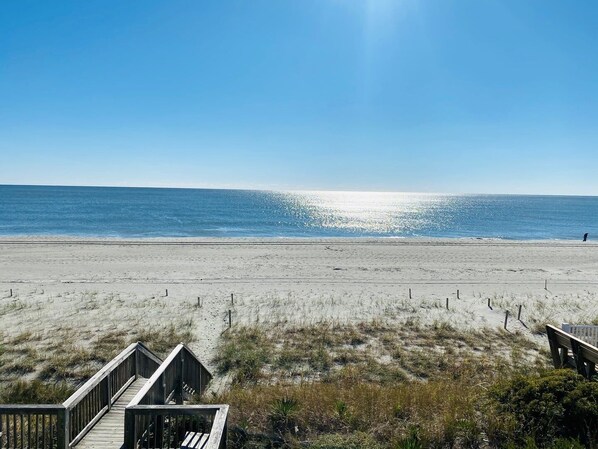 Ein Strand mit einer Holzpromenade, die zu einer Sandfläche führt, einem klaren, blauen Himmel und dem Meer im Hintergrund.