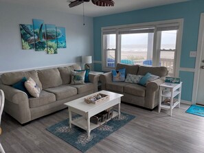 A living room with two sofas, a coffee table, and a side table with a sign. There are three wall-mounted paintings, a ceiling fan, and a view of the ocean through the windows.