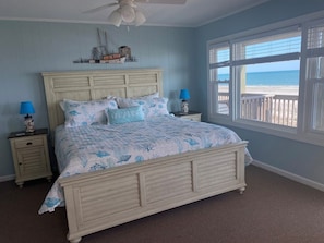 A bedroom with a bed, two nightstands, a ceiling fan, a window with a view of the beach, and a decorative ship model on the wall.