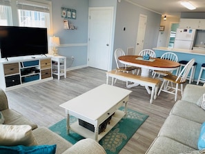 Una sala de estar con televisión, una mesa de centro blanca, un sofá y un comedor con mesa y sillas de madera.