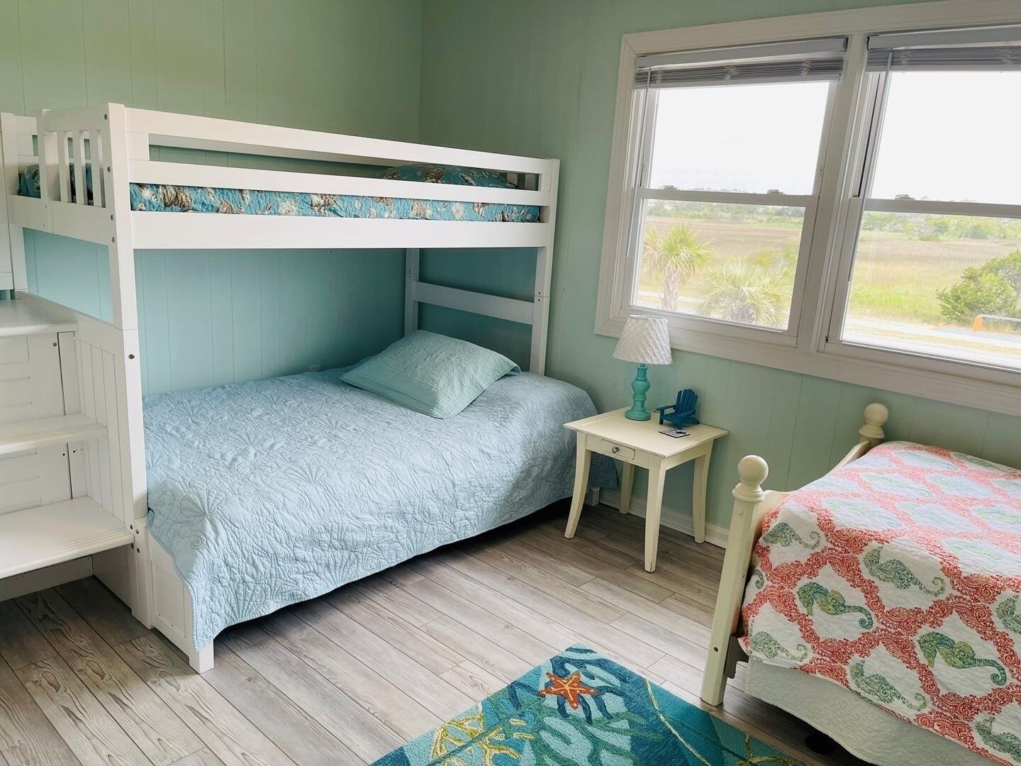 A bunk bed with a lower bunk, a bedside table with a lamp, and a window with a view of the outdoors.