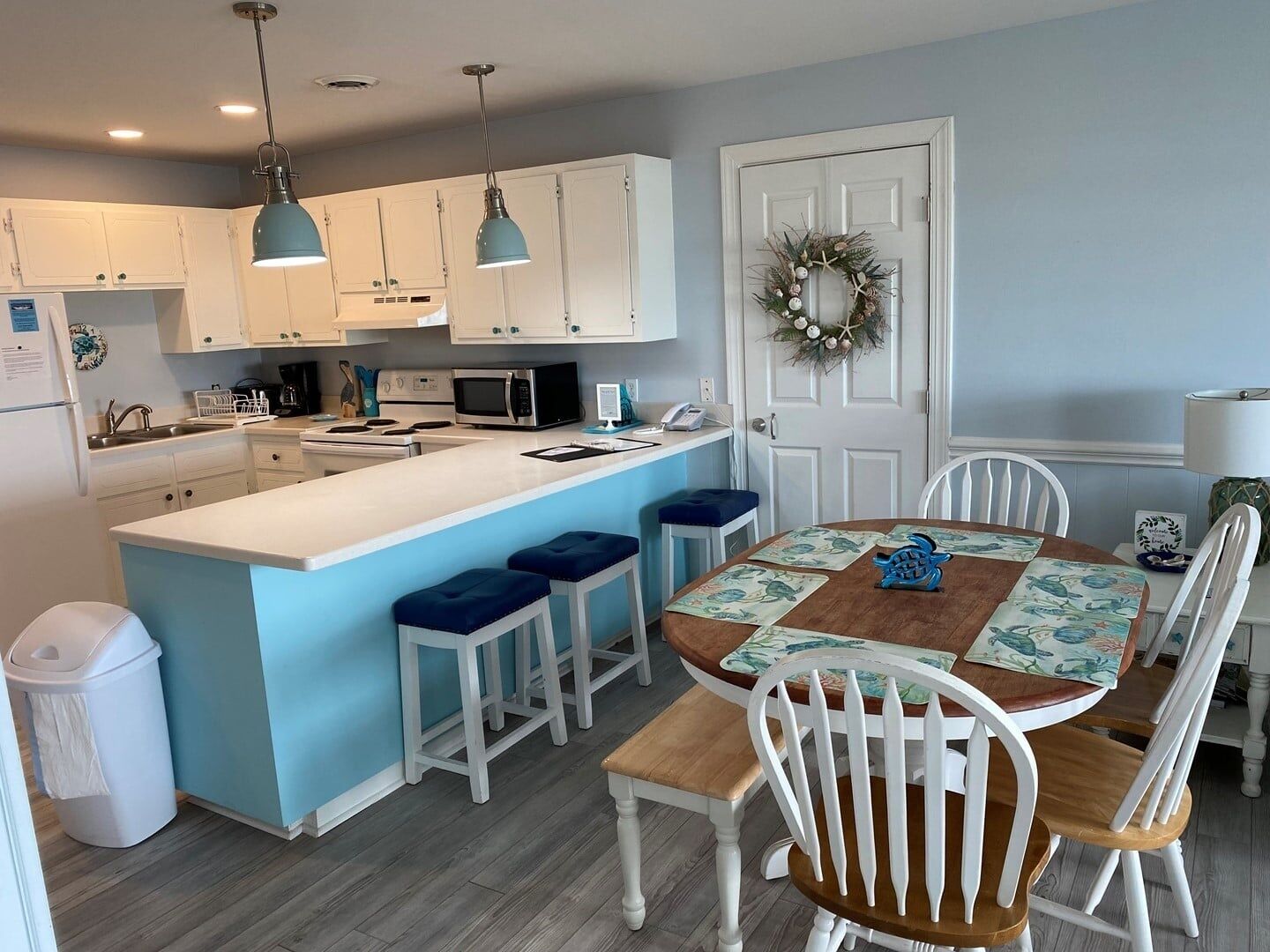 A kitchen with white cabinets, a central island with stools, a dining table with chairs, and a door with a wreath.