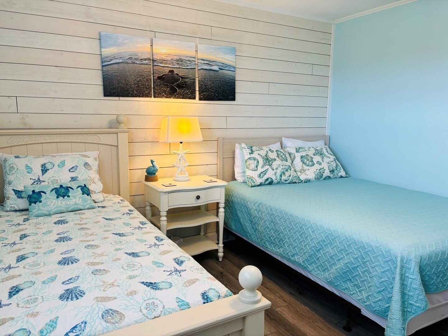 Une chambre avec deux lits, une table de chevet, une lampe et un tableau accroché au mur.