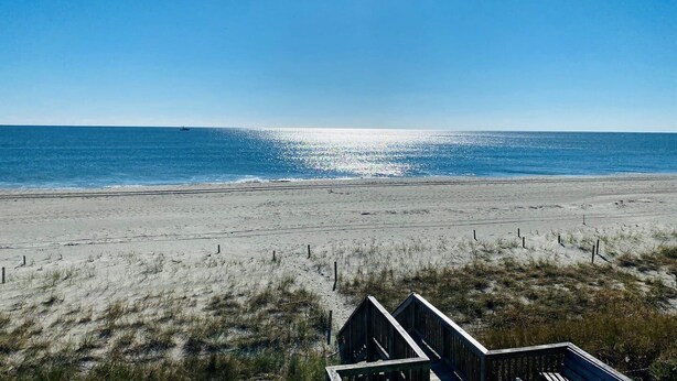 A beach with a wooden boardwalk leading to a structure, a clear blue sky, and calm ocean waters.