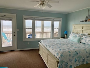 A bedroom with a bed, a ceiling fan, a window with a view of the beach, and a door leading to a balcony.